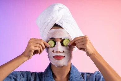 خطوات تنظيف وتفتيح وترطيب الجلد، روتين العناية وتحسين نضارة البشرة، إستخدام مكونات تجميلية, بورتريه مقرب لامراة عربية خليجية سعودية ترتدي الزي الكاجوال و منشفة الرأس تضع القناع التجميلي على الوجه مع قناع الخيار للعيون، خلفية ملونه