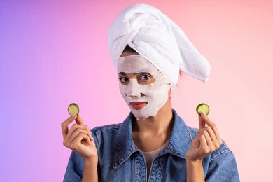 إستخدام مكونات تجميلية، مفهوم العناية والإهتمام بنضارة وتحسين البشرة ، بورتريه لمرأة عربية خليجية سعودية ترتدي الزي الكاجوال تضع منشفة الرأس تمسك بيديها قطع من الخيار، خلفية ملونه