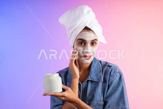 استخدام مستحضر تجميل، مفهوم العناية بنضارة وتحسين البشرة، بورتريه مقرب لامرأة عربية خليجية سعودية ترتدي منشفة الرأس و الزي الكاجوال تمسك بيدها علبة جل الترطيب،خلفية ملونه
