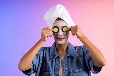 الاهتمام بالمظهر الخارجي عند النساء، مفهوم الراحة والاسترخاء، استخدام ماسكات ترطيب ومكونات تجميلية نسائية، العناية والإهتمام بنضارة وتحسين البشرة, بورتريه مقرب لامرأة عربية خليجية سعودية ترتدي الزي الكاجوال ومنشفة بيضاء على رأسها تضع شرائح من الخيار على وجهها، خلفية ملونة