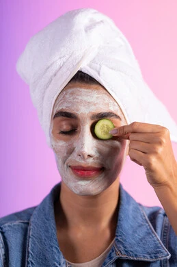 The use of moisturizing masks and women's cosmetic ingredients, the concept of care and attention to skin freshness and improvement, the concept of comfort and relaxation, a close-up portrait of a Saudi Gulf Arab woman wearing casual attire and a white towel on her head, placing slices of cucumber on her face, with a colorful background.