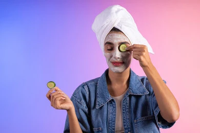 مفهوم العناية والإهتمام بنضارة وتحسين البشرة، استخدام ماسكات ترطيب ومكونات تجميلية نسائية، مفهوم الراحة والاسترخاء, بورتريه مقرب لامرأة عربية خليجية سعودية ترتدي الزي الكاجوال و منشفة بيضاء على رأسها تضع شرائح من الخيار على وجهها، خلفية ملونة