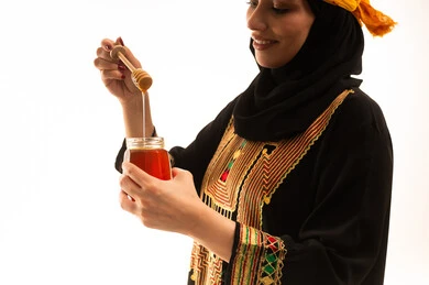 ذكرى تاسيس الدولة السعودية22 فبراير،  تعابير الفخر والسرور، إرتداء أزياء المنطقة الجنوبية، منتجات غذائية صحية ومفيدة صنعت في السعودية، بورتريه مقرب من الجانب لامراة عربية خليجية سعودية ترتدي الشيلة والسدره تحمل بيدها ملعقة مغمورة من عسل النحل الطبيعي في وعاء زجاجي، يوم بدينا1727م، خلفية بيضاء