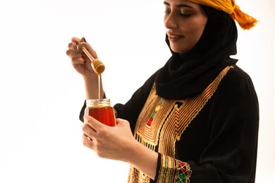 تعابير الفخر والسرور، إرتداء أزياء المنطقة الجنوبية، منتجات غذائية صحية ومفيدة صنعت في السعودية، بورتريه مقرب من الجانب لامراة عربية خليجية سعودية ترتدي الشيلة والسدره تحمل بيدها ملعقة مغمورة من عسل النحل الطبيعي في وعاء زجاجي، يوم بدينا1727م، ذكرى تاسيس الدولة السعودية22 فبراير، خلفية بيضاء