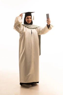 Sharing the joy of achieving the dream of success with family and friends, a portrait of a Saudi Arabian Gulf graduate wearing a graduation gown and cap, making a video call via a mobile phone, using modern technology, celebrating the completion of university studies, full-body shot, white background.