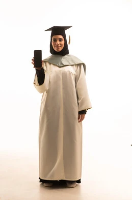 عرض شاشة سوداء فارغة، استخدام جهاز تقني حديث، إيماءات السعادة بالوصول لحلم النجاح، بورتريه لخريجة عربية خليجية سعودية محجبة ترتدي زي التخرج والقبعة تمسك بيدها هاتف محمول، الاحتفال بإنهاء الدراسة الجامعية، الجسم بالطول الكامل، خلفية بيضاء