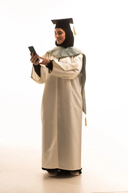 تصفح مواقع التواصل الاجتماعي عبرالجوال، التواصل مع العائلة و الأصدقاء، استخدام جهاز تقني حديث, المناسبات والاحتفالات الجامعية, بورتريه لطالبة عربية سعودية خليجية محجبة ترتدي العباءة والقبعة تمسك بيدها الهاتف المحمول, صورة الجسم بالكامل, خلفية بيضاء