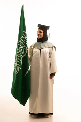 The concept of love for the homeland and belonging to it, the flags and national emblems of Arab countries, pride and honor in symbols and the flag, a portrait of a Saudi Gulf Arab graduate wearing a white graduation gown and cap, holding the Saudi flag in her hands, full-body, white background.
