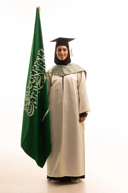 الاعتزاز والفخر بالرموز والراية، الرايات والأعلام الوطنية للدول العربية، بورتريه لخريجة عربيه خليجيه سعودية ترتدي ثوب التخرج الابيض و القبعة تمسك في يديها العلم السعودي ، مفهوم حب الوطن والإنتماء إليه، الجسم بالطول الكامل، خلفية بيضاء