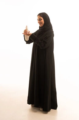 مفهوم العناية بنضارة وتحسين البشرة، بورتريه لامرأة عربية خليجية سعودية ترتدي العباءة و الحجاب تمسك علبة كريم الترطيب، استخدام مستحضر ومكون تجميلي، الاهتمام بالأناقة والمظهر الخارجي، الجسم بالطول الكامل، خلفية بيضاء