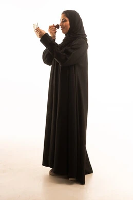 مفهوم تحسين المظهر والمكياج، التسويق لأدوات ومنتجات التجميل, بورتريه من الجانب لامراة عربية خليجية سعودية ترتدي العباءة و الحجاب تمسك فرشة المكياج بإيماءات النعومة والثقة, استعمال فرشاة أحمر الخدود ، الجسم بالطول الكامل، خلفية بيضاء