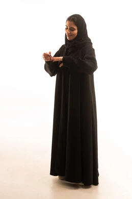 استخدام لون لطلاء الأظافر، خبيرة سعودية لمستحضرات التجميل النسائية، بورتريه لإمرأة عربية خليجية سعودية ترتدي العباءة والحجاب تحمل بيدها المانيكير، مفهوم الإهتمام بالجمال والمظهر الخارجي، صالون تجميلي نسائي، صورة الجسم بالكامل، خلفية بيضاء