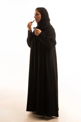 The concept of adornment and beautification, promoting cosmetic products and makeup, using cosmetic products, taking care of beauty and appearance, a portrait of a Saudi Arabian Gulf woman wearing a black abaya and applying lipstick, putting on makeup, full-body, white background.