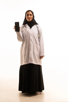 عرض شاشة سوداء فارغة عبر الجوال، خدمات و تطبيقات طبية عبر الهاتف، تلبية مطالب الرعاية الصحية عن بعد باستخدام التقنيات الحديثه، بورتريه لطبيبة عربية خليجية سعودية ترتدي العباءة و المعطف الطبي تضع السماعة الطبية تحمل الهاتف المحمول، الجسم بالطول الكامل، خلفية بيضاء