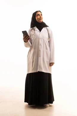 النظر الى شيء ما ، تقديم الخدمات للمرضى بكل كفاءه, اجراء الاستشارات الطبيه عن بعد, استخدام الجوال في البحث والتطوير، بورتريه لطبيبة عربية خليجية سعودية ترتدي المعطف الابيض تحمل الهاتف المحمول ،  الجسم بالطول الكامل، خلفية بيضاء