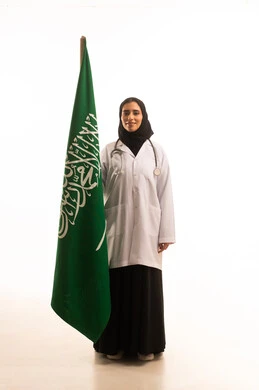 ذكرى توحيد المملكة العربية السعودية، بورتريه لطبيبة عربية سعودية تلبس رداء وسماعة الطبيب تمسك بيديها علم السعودية تحتفل باليوم الوطني بفخر وعزة , العلم السعودي راية التوحيد والاسلام , الجسم بالطول الكامل، خلفية بيضاء