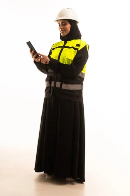 الدردشة مع العائلة والأصدقاء، تصفح مواقع التواصل الاجتماعي، استخدام جهاز تقني حديث متطور، مفهوم الهندسة والإنشاء المعماري, بورتريه لمهندسة عربية خليجية سعودية محجبة ترتدي العباءة والسترة و خوذة الحماية تمسك بيدها الهاتف المحمول، صورة الجسم بالطول الكامل، خلفية بيضاء