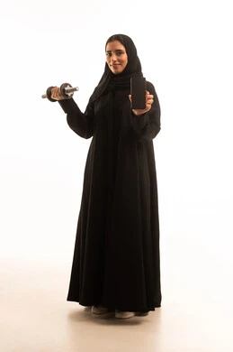 Maintaining health, physical fitness, and physical agility, displaying a blank black screen on a mobile phone, exercising and lifting weights with dumbbells, a portrait of a young Arab Gulf Saudi woman wearing a hijab and an abaya, holding heavy weights in her hand, full-body shot, white background.