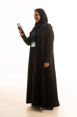تصفح مواقع التواصل الاجتماعي عبر الهاتف المحمول, استخدام جهاز تقني حديث متطور, بورتريه لامرأة عربية خليجية سعودية محجبة ترتدي العباءة تضع بطاقة بيضاء فارغه بشريط أزرق حول رقبتها، موك اب ابيض، التواصل مع الاهل والاصدقاء، صورة الجسم بالطول الكامل, خلفيه بيضاء