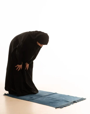 The five pillars of Islam, the concept of worship and drawing closer to God, performing the obligatory prayer, a portrait of a young Arab Gulf Saudi woman wearing an abaya and hijab standing on a designated prayer rug in a bowing position, full-body image, white background.