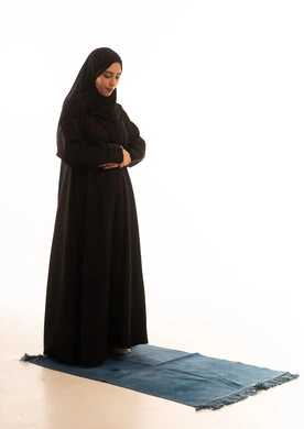 أداء الفروض على وقتها، ايماءات تدل على الخشوع في الصلاة, التقرب الى الله من خلال الإجتهاد في العبادة والحفاظ على الصلوات، بورتريه لإمرأة عربية سعودية خليجية ترتدي الحجاب والعباءة تقف على سجادة الصلاة, صورة الجسم بالطول الكامل, خلفية بيضاء