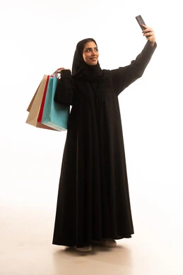 التسوق من المولات التجارية، مفهوم التصوير والشهرة عبر برامج التواصل الاجتماعي، بورتريه لامراة عربية خليجية سعودية ترتدي عباءة سوداء تحمل بيدها أكياس التسوق الملونه، التقاط صورة سيلفي عبر الجوال، الجسم بالطول الكامل، خلفية بيضاء