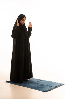 Gestures indicating humility in prayer, performing obligatory prayers on time, drawing closer to God through diligence in worship and maintaining prayers, a portrait of a Saudi Arabian Gulf woman wearing a hijab and abaya standing on a prayer rug, full-body image, white background.