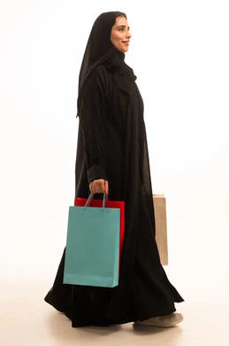 الأثر الإيجابي للتسوق على السيدات، الاستفادة من العروض الموسمية والتخفيضات، شراء المستلزمات والتبضع من المولات التجارية، بورتريه من الجانب لسيده عربية خليجية سعودية ترتدي العباءه والحجاب تحمل بيدها أكياس التسوق الملونه، الجسم بالطول الكامل، خلفية بيضاء