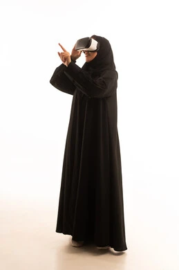 The enjoyment of watching through virtual reality glasses, trying to click on something, a side portrait of a veiled Arab Gulf Saudi woman wearing an abaya and VR glasses, immersion and simulation of the imaginary world, a young Saudi woman raising her index finger and pointing to a specific place, full-body shot, white background.