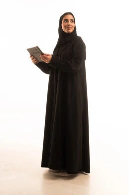 النظر الى شيء ما، تصفح مواقع ووسائل التواصل الاجتماعي، دمج التقنية والتكنولوجيا الحديثة في الحياة اليومية، بورتريه لامرأة عربية خليجية سعودية ترتدي الحجاب والعباءة تقوم بالنقر على التابلت، شابة سعودية تعمل على الجهاز اللوحي، صورة لكامل الجسم، خلفية بيضاء