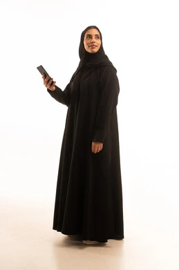 النظر الى شيء ما، تصفح وسائل التواصل الاجتماعي باستخدام الجوال، بورتريه لإمرأة عربية خليجية سعودية مبتسمة ترتدي حجاب وعباءة سوداء تمسك بيديها الهاتف المحمول، الدردشة والتواصل مع التواصل مع الأصدقاء اونلاين، صورة الجسم بالكامل، خلفية بيضاء