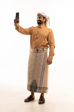 التقاط صور السيلفي التذكارية، مفهوم التقنيات والأجهزة الحديثة، بورتريه لرجل عربي يمني يرتدي القميص والازار مع العمامة يحمل بيده الجوال، صورة الجسم بالطول الكامل، خلفية بيضاء