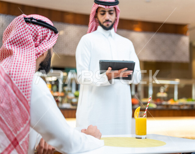 رجل عربي سعودي خليجي يجلس في مقهى, شاب يطلب الطعام من النادل في مطعم, تسجيل طلبات الطعام في جهاز التابلت, قضاء وقت ممتع وهادئ