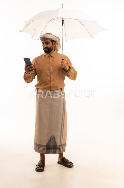 Browsing social media on a mobile phone, a portrait of an Arab Yemeni man wearing a shirt and a sarong with a turban holding an umbrella in his hand, protection from rain in winter, full-body, white background.