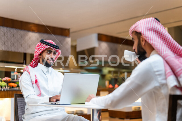 Two Saudi Arabian Gulf colleagues sitting in a cafe, discussing work tasks in the restaurant, a work dialogue between two Saudi Gulf men in a restaurant, completing work tasks on the computer in the cafe, acquaintance and building social relationships, fr