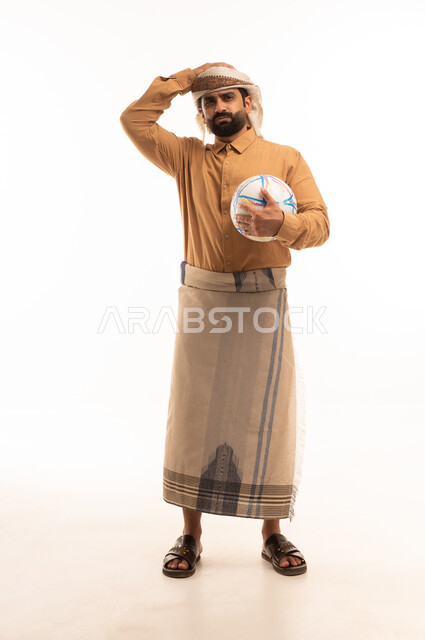 Gestures of loss and failure, watching and following a football match, a portrait of a Yemeni Arab man wearing a shirt and a sarong holding a football and placing his hand on his head with expressions of sadness and interaction with the national team, different sports clubs, white background.
