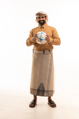 النظر الى الكاميرا بايماءات السرور والسعادة، بورتريه لرجل عربي يمني يرتدي القميص والازار مع العمامة يحمل كرة قدم يقوم بمشاهدة المباريات، قضاء وقت ممتع في المنزل، الشغف في متابعه الفرق وتشجيع المنتخبات، خلفية بيضاء