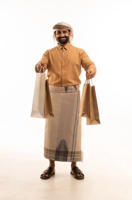 النظر الى الكاميرا بتعابير السرور، التبضع وأخذ الحاجات اللازمة, المشتريات التجارية والهدايا, الاستفادة من الحسومات والتصفيات, بورتريه لرجل عربي يمني يرتدي القميص والازار مع العمامة يمسك بيده أكياس ورقية ملونة, الجسم بالطول الكامل, خلفية بيضاء