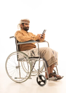 متابعة مواقع التواصل الاجتماعي عبر الهاتف المحمول، مشاهدة الأفلام والمسلسلات الممتعة، التواصل مع الأهل والأصدقاء، استخدام التكنولوجيا و التقنيات الحديثة,  بورتريه لرجل عربي يمني يرتدي القميص والازار مع العمامة يجلس على كرسي متحرك يحمل بيده موبايل، خلفية بيضاء