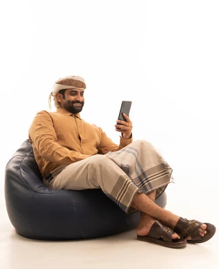 The use of modern technological devices for entertainment and leisure, browsing social media, a portrait of an Arab Yemeni man wearing a shirt and a sarong with a turban sitting on a chair holding a mobile phone in his hand, white background.