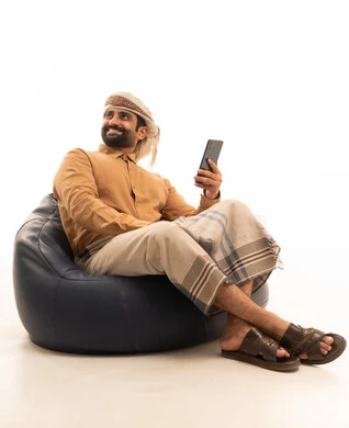 Looking at something, browsing social media, using modern technological devices for entertainment and leisure, a portrait of an Arab Yemeni man wearing a shirt and a waist wrap with a turban sitting on a chair holding a mobile phone in his hand, white background.