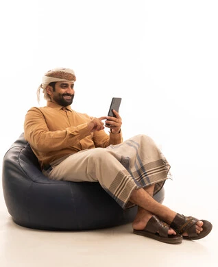 تصفح وسائل التواصل الاجتماعي , استخدام الأجهزة التقنية الحديثة للتسلية و الترفيه ,بورتريه لرجل عربي يمني يرتدي القميص والازار مع العمامة يجلس على كرسي بين باج يمسك يمسك الهاتف المحمول بيده , خلفية بيضاء