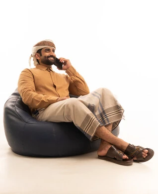 Making a phone call via mobile, gestures of happiness and joy, a portrait of a Yemeni Arab man wearing a shirt and a sarong with a turban sitting on a chair holding a mobile phone in his hands, white background.