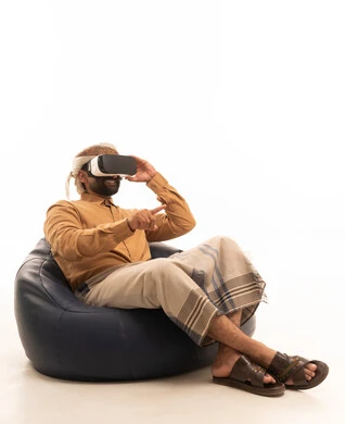 The enjoyment and entertainment of watching through virtual reality glasses, pointing to something, immersion and simulation of the imaginary world, a portrait of an Arab Yemeni man wearing a shirt and a waist wrap with a turban sitting on a chair between bags wearing 3D glasses (VR), technological and technical development, full-body, white background.