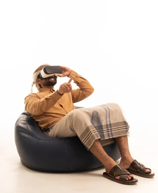 الاشارة الى شيء ما، المتعة والتسلية بالمشاهدة عبر نظارة الواقع الافتراضي، الاندماج ومحاكاة العالم الخيالي، بورتريه لرجل عربي يمني يرتدي القميص والازار مع العمامة يجلس على كرسي بين باج يضع نظارة ثلاثية الأبعاد (الفي ار), التطور التقني والتكنولوجي ، خلفية بيضاء