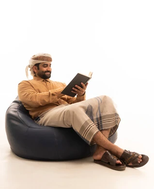 The concept of literacy and learning, spending leisure time reading, future planning for goals and aspirations, a portrait of a Yemeni Arab man wearing a shirt, izar, and turban, sitting on a beanbag chair, holding a book which he is reading intently, white background