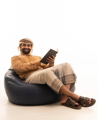 التخطيط المستقبلي للأهداف والطموحات، قضاء وقت الفراغ بالقراءة والمطالعة، مفهوم محو الأمية والتعلم, بورتريه لرجل عربي يمني يرتدي القميص والازار مع العمامة يجلس على كرسي بين باج ويمسك بيديه كتاب، تعابير السعادة و السرور، خلفية بيضاء
