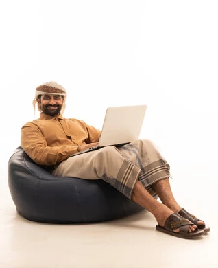 إنجاز مهام العمل عن بعد، جهاز تقني حديث ومتطور، تعابير السعادة و السرور، ورتريه لرجل عربي يمني يرتدي القميص والازار مع العمامة يجلس على كرسي بين باج يقوم باستخدام الحاسوب المحمول، خلفية بيضاء