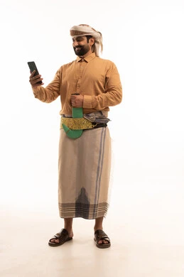 اجراء مكالمه فيديو، استخدام التقنيات والتكنولوجيا الحديثة بالحياة اليومية، الدردشة والتحدث مع الأصدقاء،تصفح مواقع وتطبيقات الانترنت، بورتريه لرجل عربي يمني يرتدي القميص والازار مع العمامة والجنبية يمسك الجوال بيده، الجسم بالطول الكامل، خلفية بيضاء