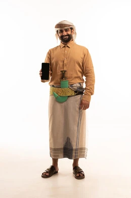 A blank black screen displayed on a mobile phone, depicting the use of modern technology in daily life, browsing websites and internet applications. The image shows a portrait of a Yemeni Arab man wearing a shirt and izar (traditional Yemeni sarong), turban, and jambiya (traditional dagger), holding a mobile phone. The image is in full length and has a white background.